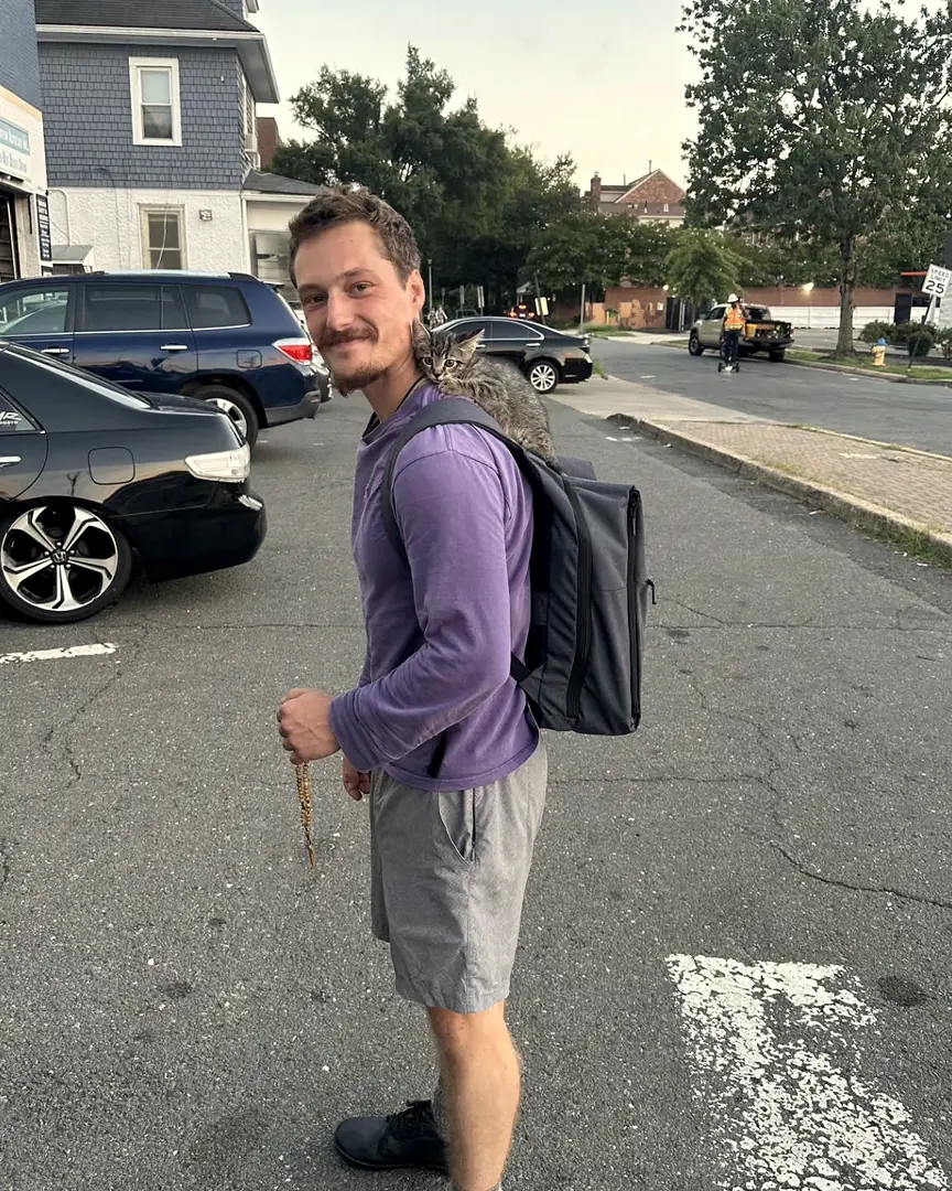 John Stamm walking with his backpack and rosary, a kitten riding on his shoulder
